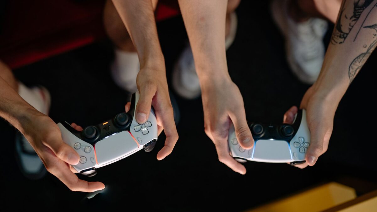 Vue de dessus de deux paires de mains tenant chacune une manette PlayStation blanche dans une pièce sombre, avec des baskets en bas de l'image et un tatouage sur un bras.