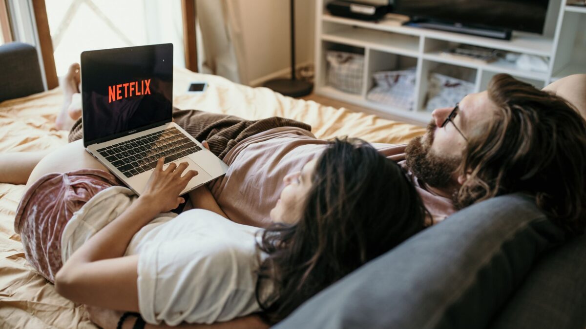 Un couple est allongé sur un lit aux draps beige, un homme barbu à lunettes et une femme aux cheveux sombres partagent un ordinateur portable affichant le logo rouge NETFLIX ; une télévision et une étagère se trouvent en arrière-plan.