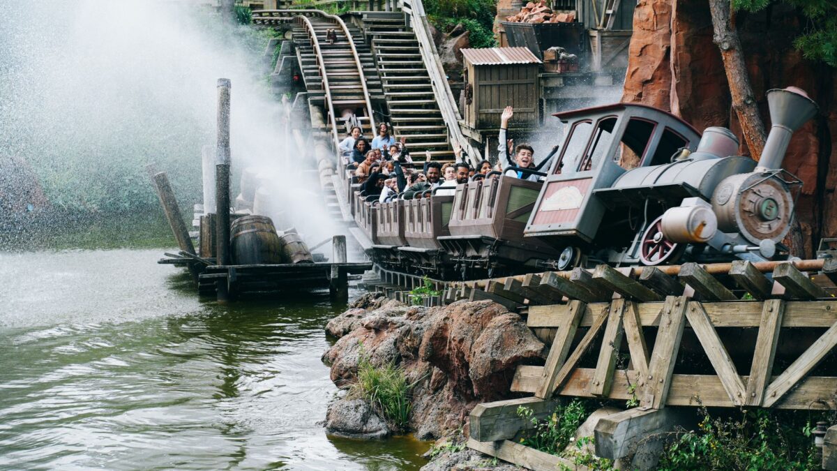 Un train à vapeur de type Big Thunder Mountain roule sur des rails en bois au bord d'un cours d'eau, éclaboussant l'eau. Des passagers lèvent les bras sur les wagons, devant des rochers rouges et de la végétation.