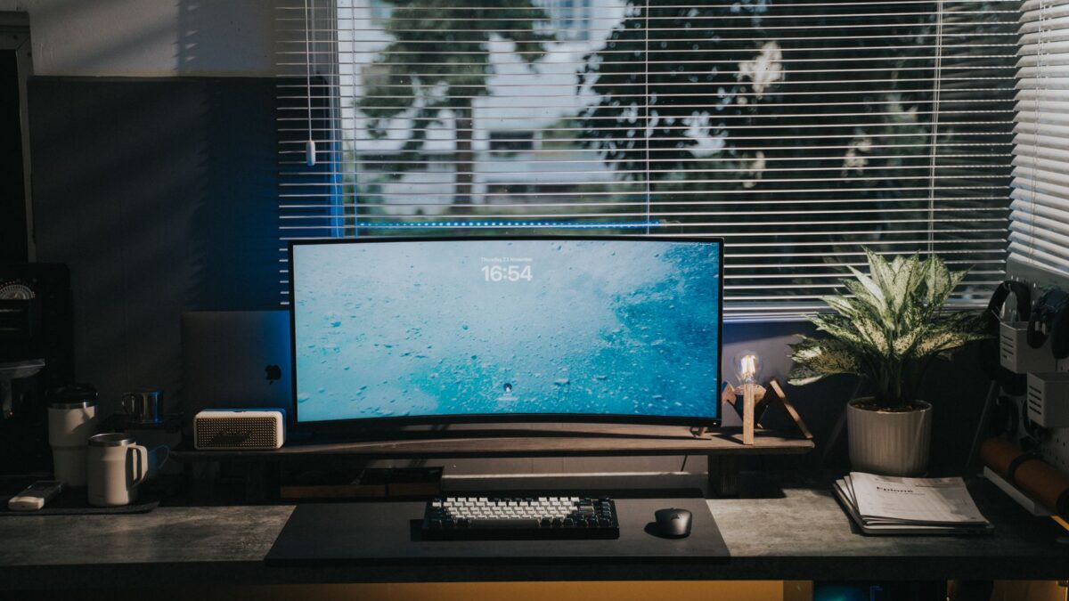 Bureau avec un grand écran incurvé affichant un fond bleu marin et 16:54, sur un support en bois, près d'un clavier, d'une souris, de tasses, d'une plante verte et d'un ordinateur portable.