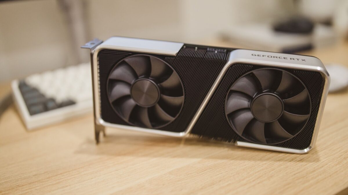Une carte graphique GeForce RTX à deux ventilateurs posée sur un bureau en bois, avec un clavier et un écran flou en arrière-plan.