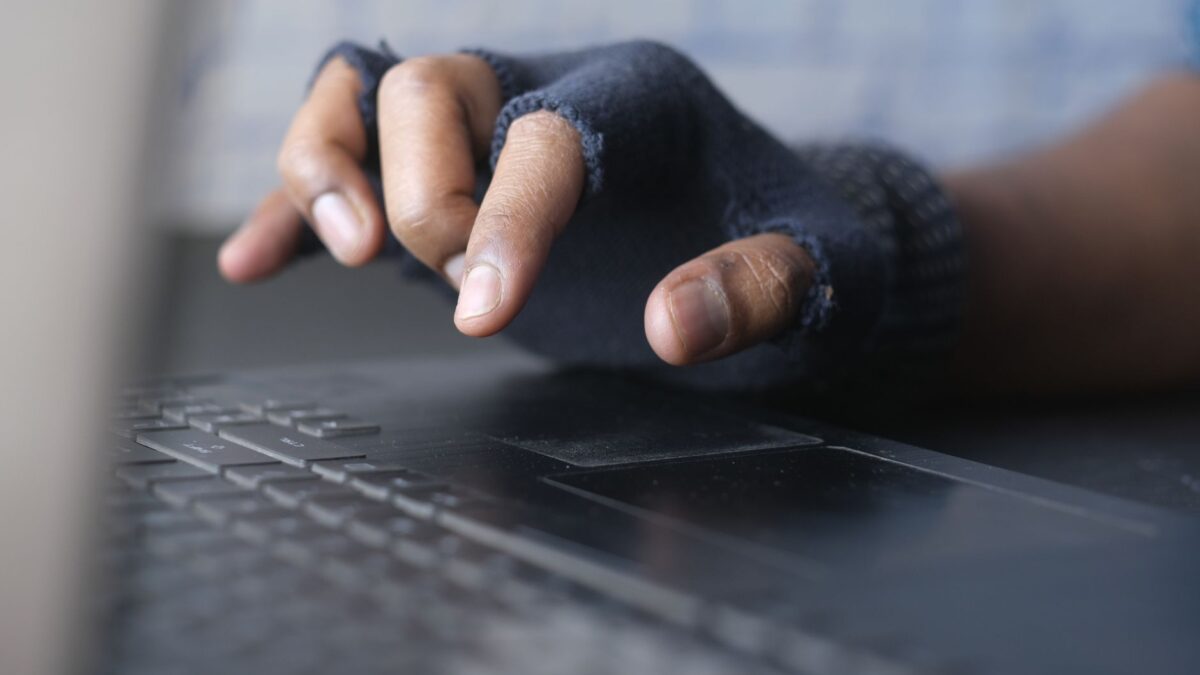 Gros plan d'une main gantée, aux doigts découverts en bleu foncé, posée au-dessus d'un clavier d'ordinateur portable sombre, sur fond clair flou.