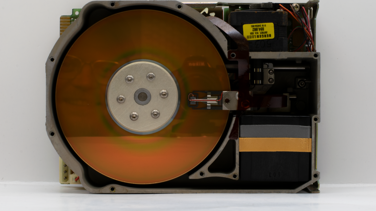 Vue en coupe d'un disque dur ouvert montrant un plateau magnétique orangé, un bras de lecture avec tête et une carte électronique ancienne à étiquette jaune sur fond blanc.