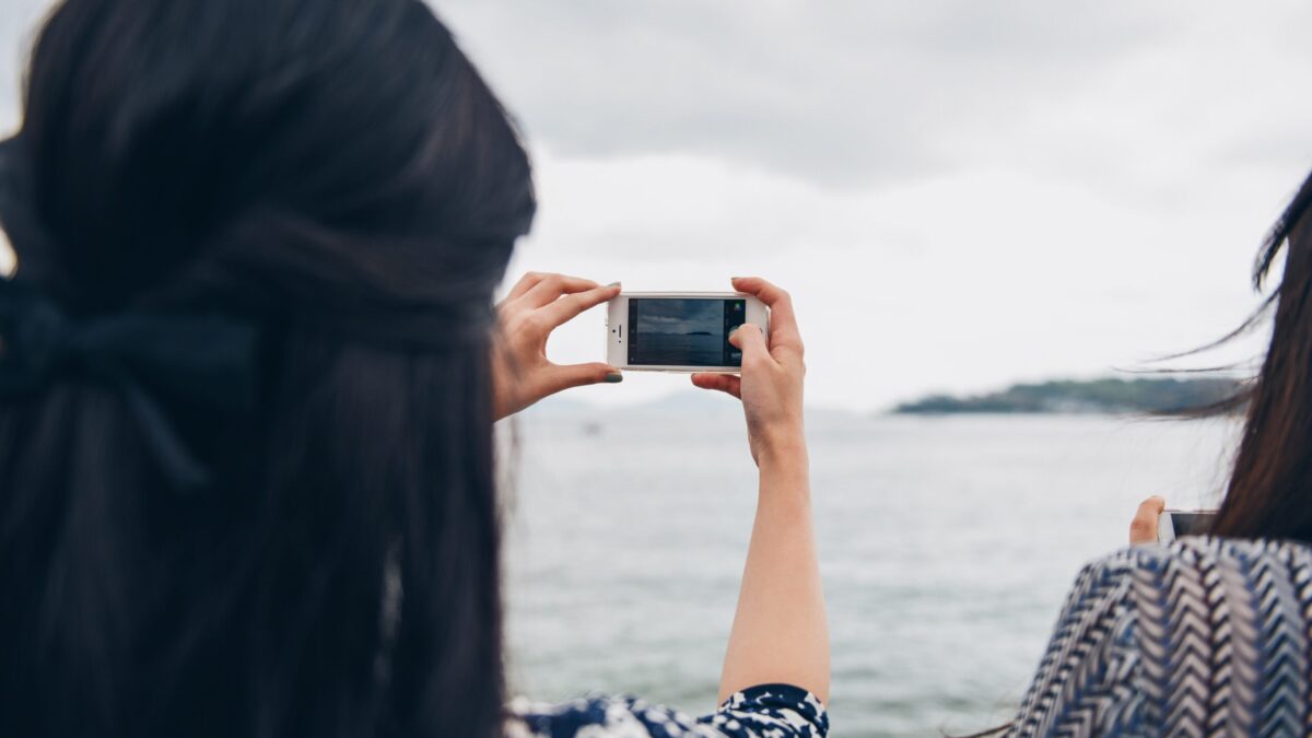 Personne prenant une photo avec un smartphone au bord de la mer, paysage aquatique et ciel nuageux en arrière-plan.