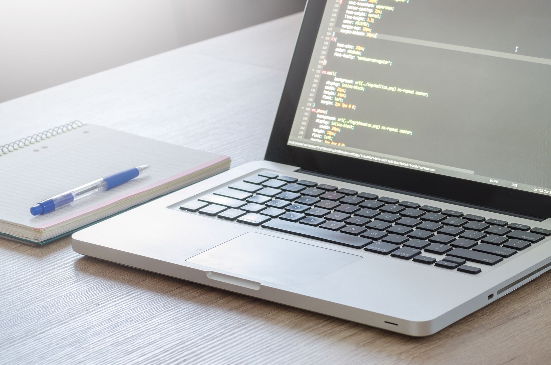 An open laptop displaying lines of code on its screen on a wooden desk next to a notebook.