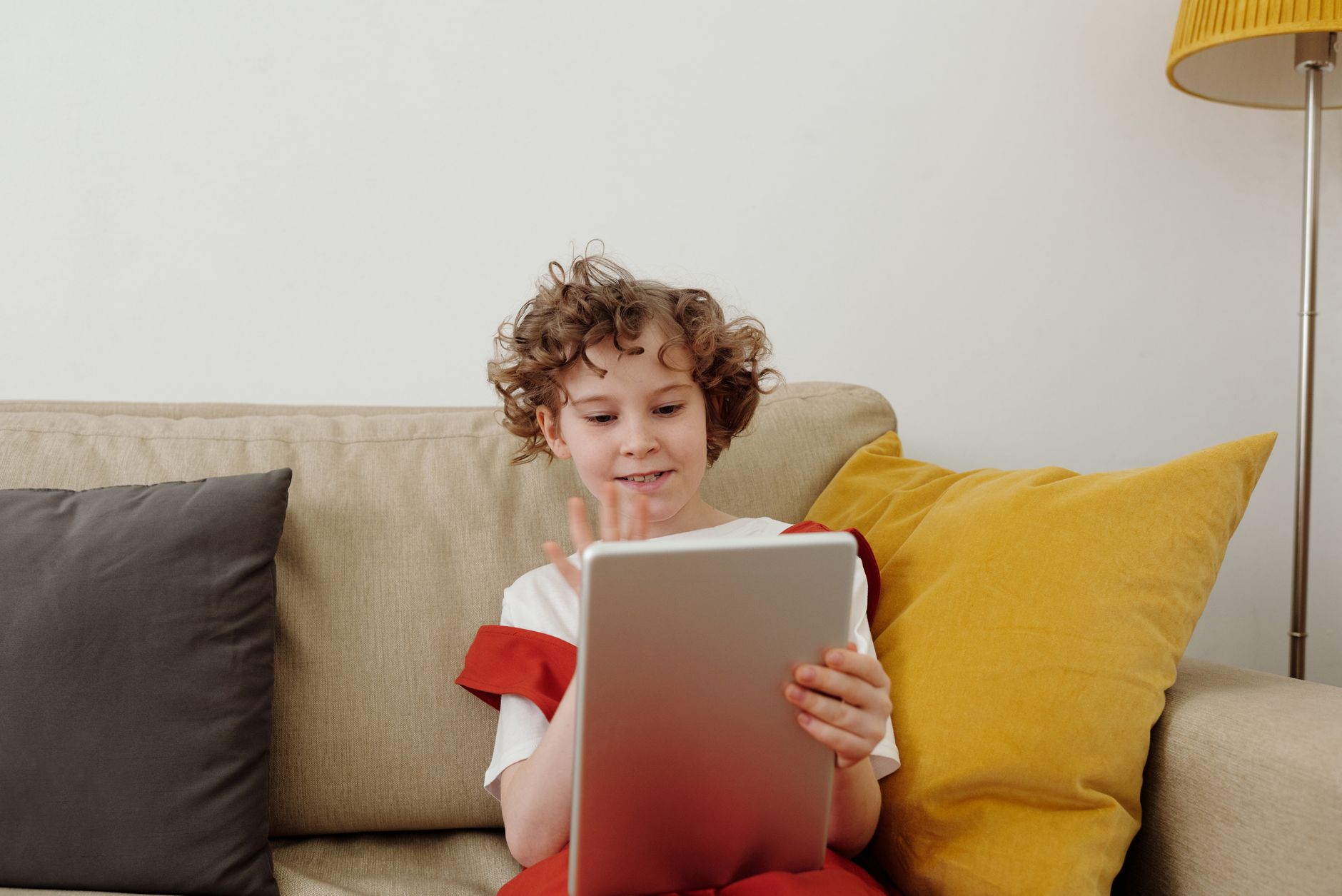 Child with curly hair sitting on a beige sofa, holding a silver tablet and waving a hand at it, with yellow and grey cushions nearby.