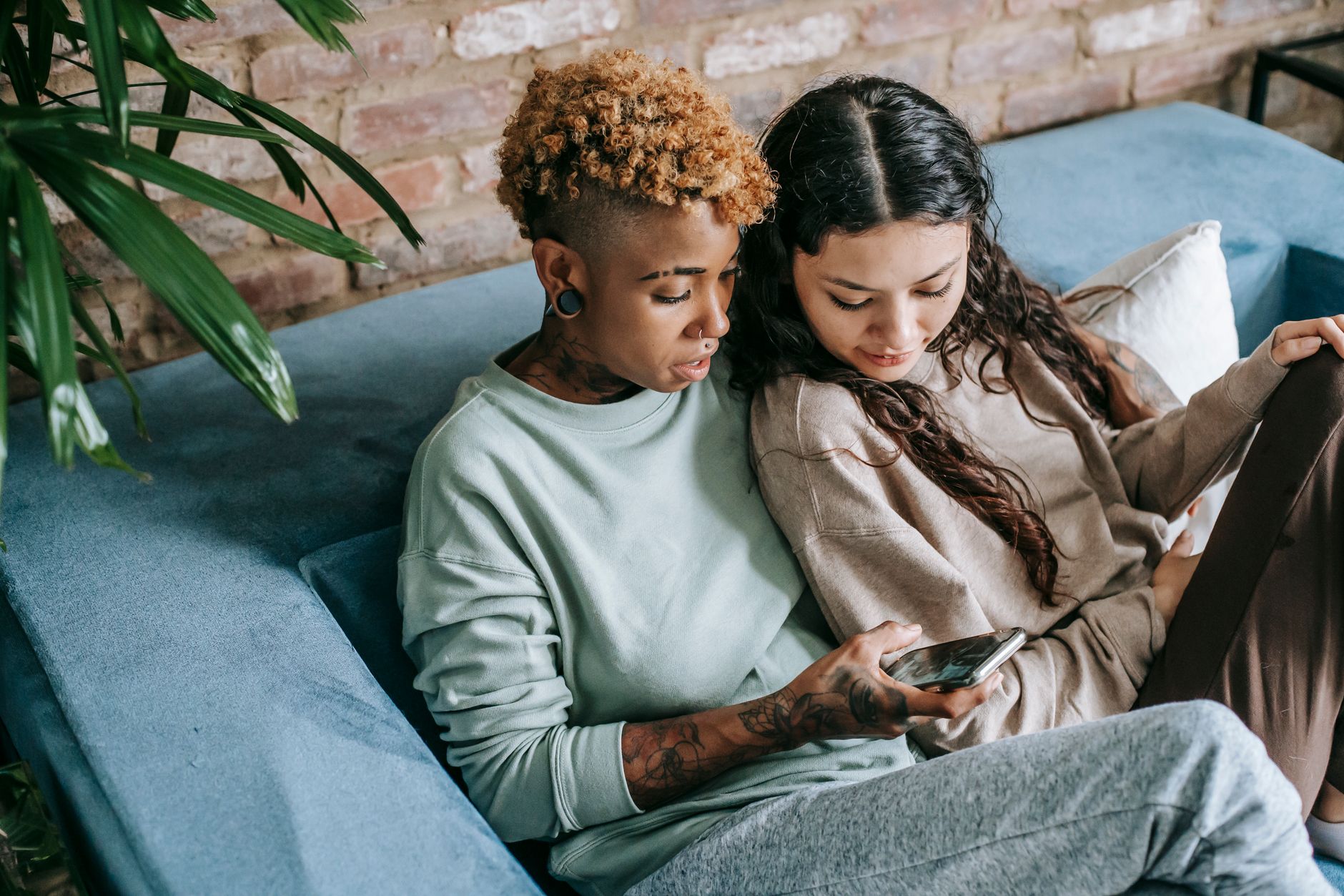 Deux femmes assises sur un canapé bleu regardent ensemble un smartphone, devant un mur de briques et une plante verte.