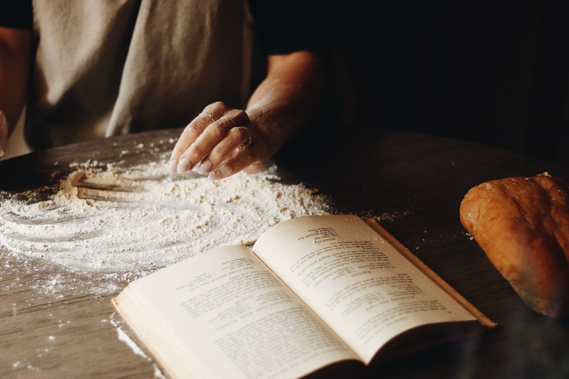Flour-dusted hand on a wooden table beside an open printed recipe book and a loaf of bread.