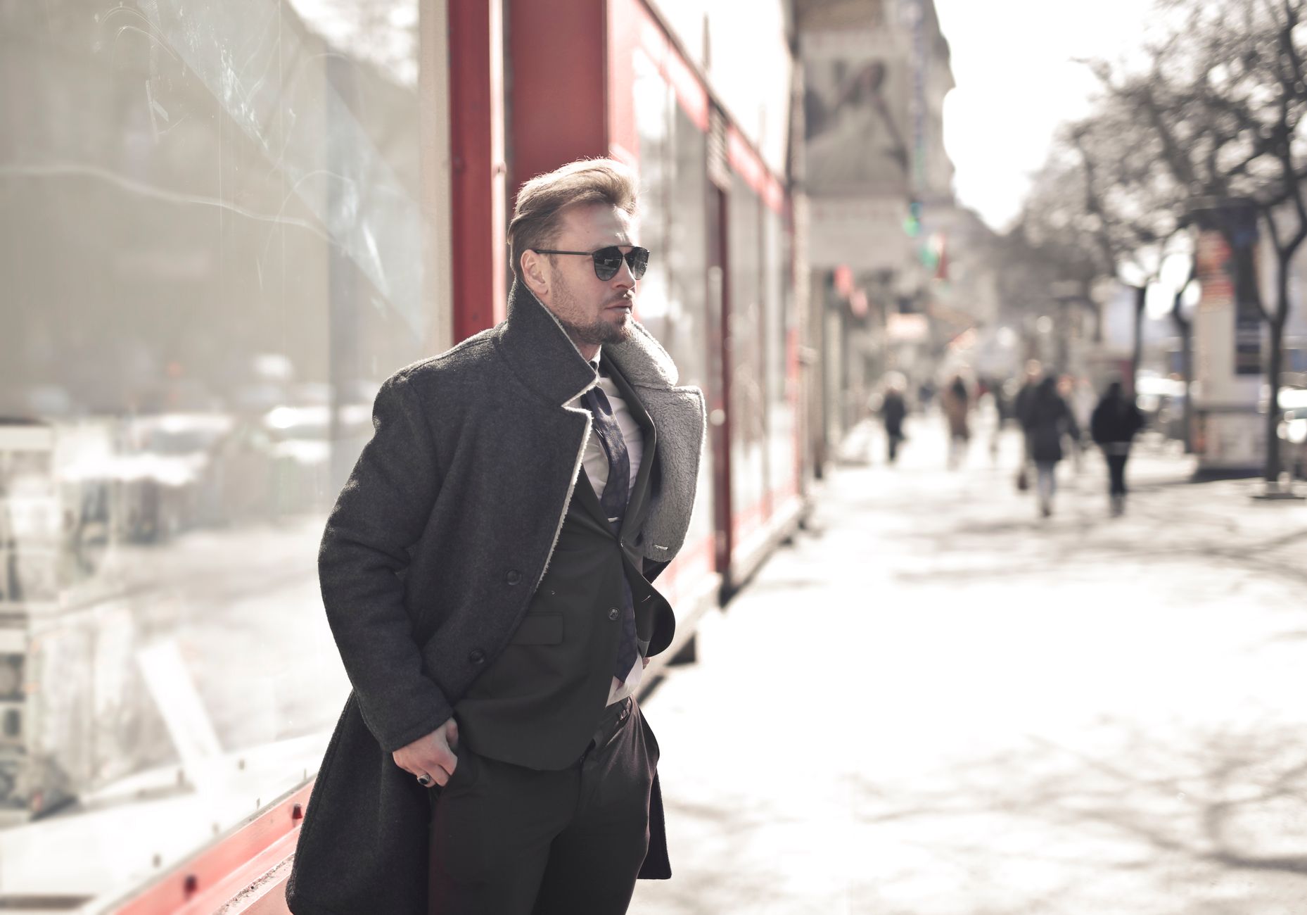 Man in sunglasses and a gray wool coat stands on a sunny city sidewalk while blurred pedestrians walk behind him.