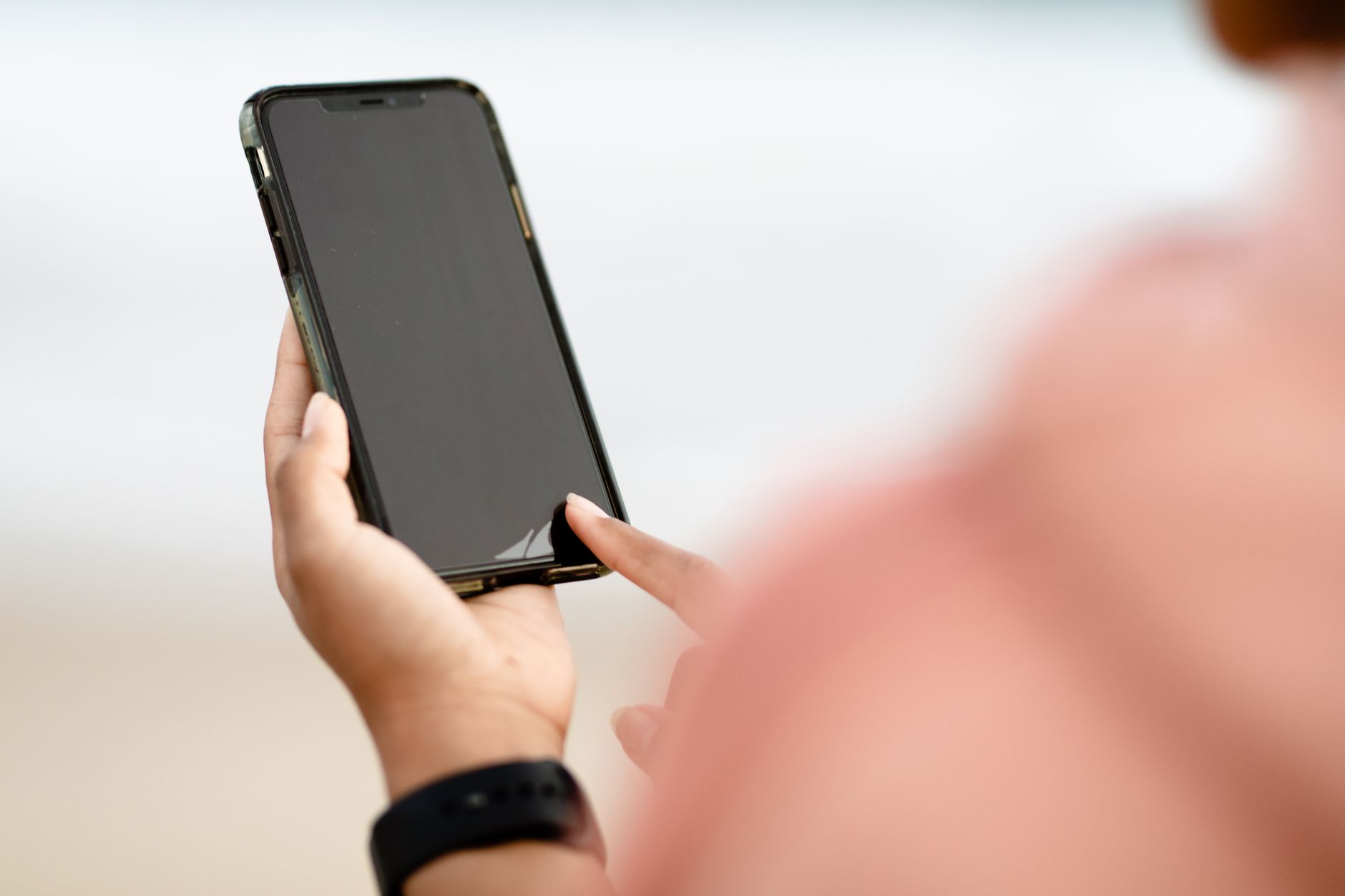 Person holding a smartphone with a dark screen and touching it with one finger, wearing a black wristband against a blurred light background.