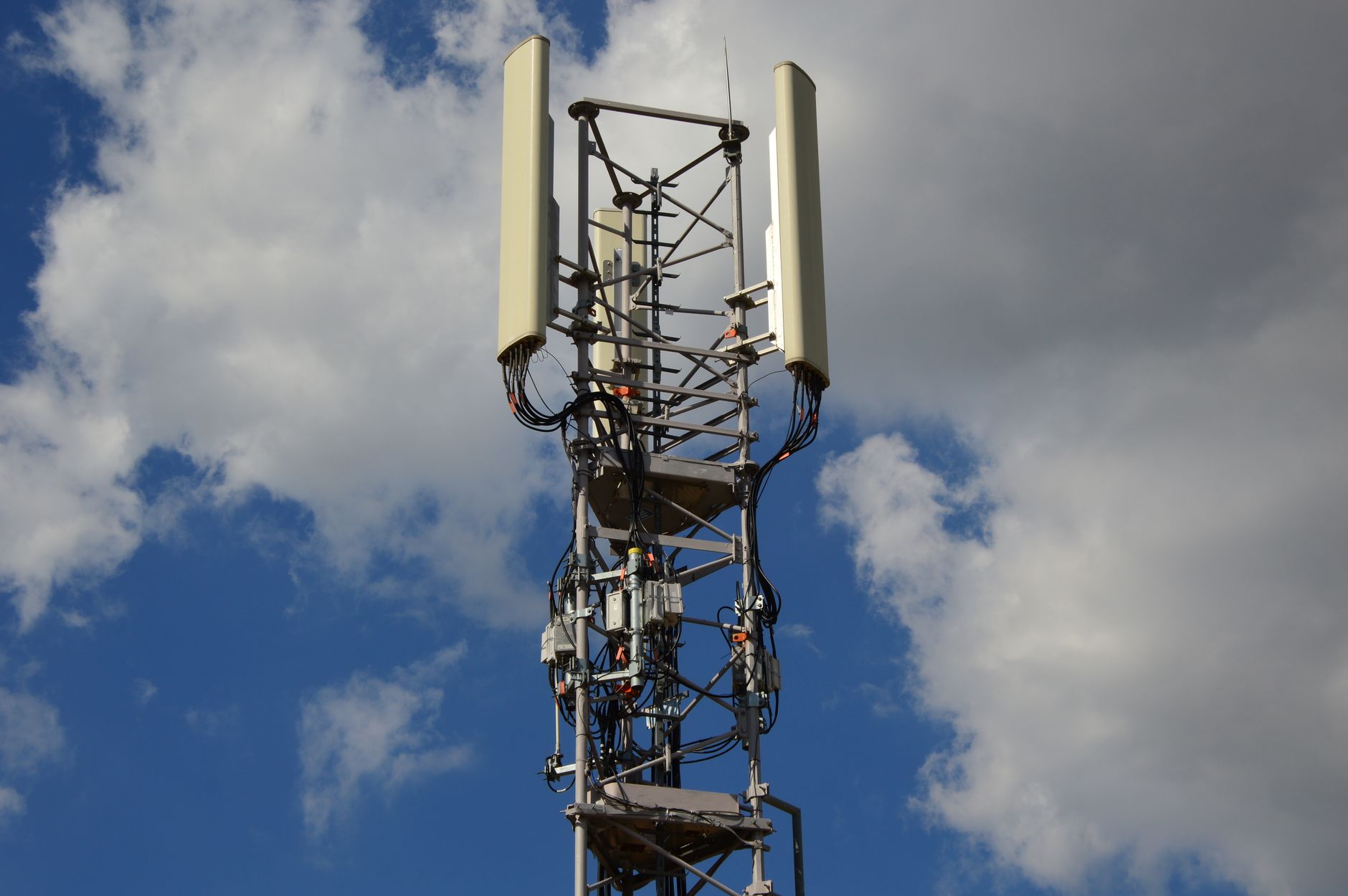 Top of a lattice cell tower with panel antennas, equipment boxes and cables against a blue sky with white clouds.